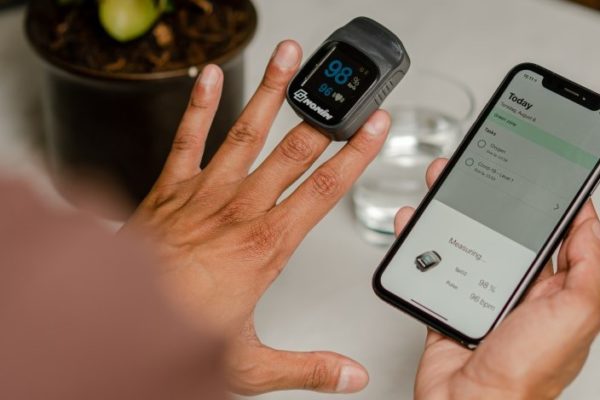 man wears a pulse oximetry device on his left middle finger to measure oxygen saturation, while holding a smartphone in his right hand