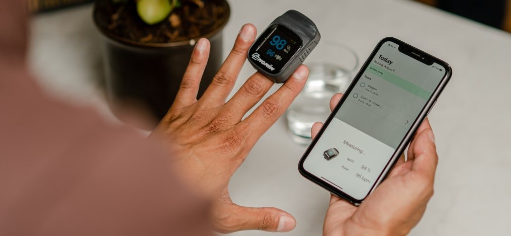 man wears a pulse oximetry device on his left middle finger to measure oxygen saturation, while holding a smartphone in his right hand